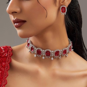 May include: A dazzling jewelry set featuring a ruby red necklace and matching earrings. The necklace is a choker style with square-cut red stones, surrounded by a silver or white metal setting. The earrings have a similar design, with a single red stone.