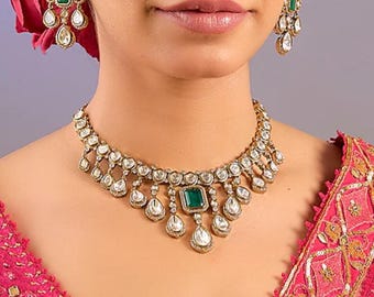 May include: A gold necklace with a large emerald green stone in the center and smaller clear stones surrounding it. The necklace has a teardrop design with smaller teardrop stones hanging from the center stone. The necklace is worn by a person with a pink and gold embroidered blouse.