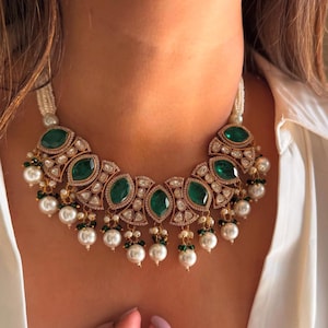 May include: A gold-toned necklace featuring large, oval emerald-green gemstones and smaller clear stones. The necklace has a pearl strand and dangling pearl and green bead accents. The design is symmetrical and ornate, with a classic, elegant style.