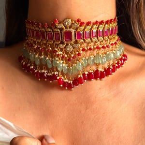 Puede incluir: Un collar gargantilla dorado con piedras preciosas rojas y verdes. El collar presenta una serie de piedras rojas rectangulares, intercaladas con pequeñas cuentas rojas y verdes. Las cuentas rojas colgantes y pequeñas perlas blancas crean un efecto en capas.