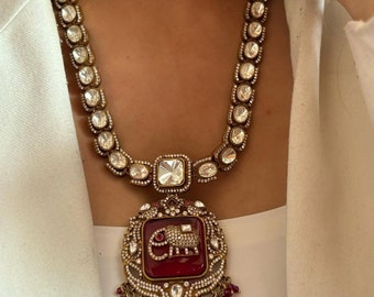 Op de afbeelding: Een gouden ketting met een grote rode hanger met een olifantendesign. De hanger is omringd door kleinere witte stenen en heeft een franje van rode kralen.