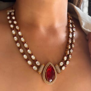 May include: A gold necklace with a large red gemstone pendant. The necklace is made of two strands of small white and red stones.