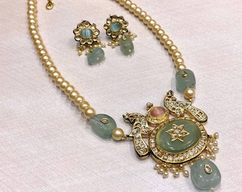 Op de afbeelding: Een groene en gouden ketting met een grote, ovale hanger met een stervormig ontwerp en twee pauwen. De hanger is omgeven door kleinere stenen en parels. De ketting is geregen op een witte parelstreng.