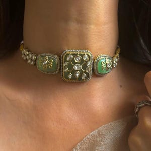 May include: A gold choker necklace with green and clear stones. The necklace has a square-shaped center piece with a floral design.