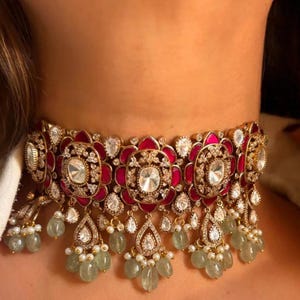 Puede incluir: Un collar gargantilla dorado con un diseño floral. El collar presenta grandes gemas transparentes, detalles en rojo y cuentas verdes colgantes. Es una joya ornamentada.