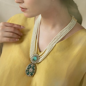 Op de afbeelding: Een meerstrengs parelketting met een grote, ovale hanger. De hanger is gemaakt van goud en heeft een groene steen die omringd is door kleinere stenen. De ketting wordt gedragen door een persoon met een geel shirt.