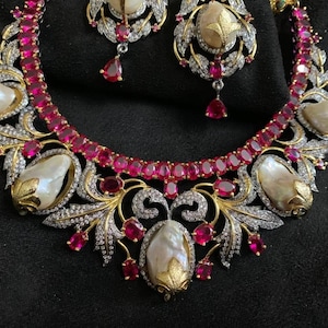 以下が含まれることがあります： ピンクの宝石と白い真珠のようなアクセントが施された、花柄の金のネックレスとイヤリングのセットです。