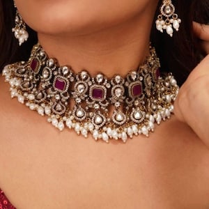 Pode incluir: Um close-up de um conjunto de colar e brincos. O colar apresenta pedras preciosas quadradas, vermelho escuro, rodeadas por pedras transparentes e pequenas pérolas brancas. Os brincos têm um design semelhante, com pérolas brancas.