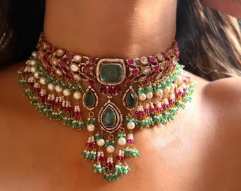 May include: A gold choker necklace with emerald green and ruby red gemstones. The necklace features a central square emerald, teardrop-shaped emerald pendants, and dangling pearl and gemstone clusters. The design includes white pearls and small red and green beads.