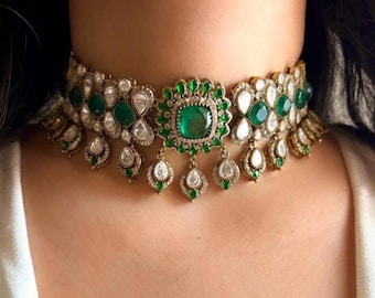 Op de afbeelding: Een goudkleurige choker ketting met smaragdgroene en heldere edelstenen. De ketting heeft een centrale vierkante smaragd, omringd door kleinere groene stenen en druppelvormige hangers. Het ontwerp is sierlijk en gedetailleerd.
