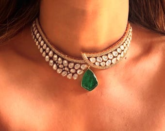 May include: A gold choker necklace with a large emerald green gemstone pendant. The necklace is adorned with small, clear gemstones.