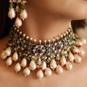 May include: An elaborate necklace and earring set. The dark metal base is decorated with clear and red gemstones, small pearls, and large teardrop pearls. The jewellery features intricate floral patterns and green accents.