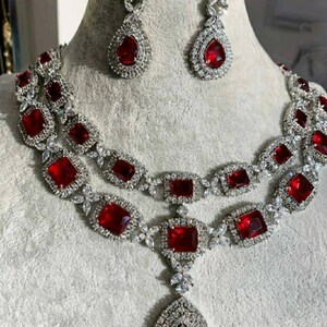 May include: A silver-toned jewelry set featuring a necklace and matching earrings. The necklace has two strands with square and rectangular red gemstones, and a large teardrop pendant. The earrings have a teardrop-shaped red gemstone.