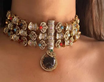 Puede incluir: Un collar dorado con múltiples capas de detalles ornamentados. El collar presenta un colgante central con una piedra oscura grande y está adornado con acentos coloridos y adornos brillantes. El collar está diseñado para ser usado alrededor del cuello.