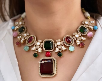May include: A gold necklace with a large red gemstone pendant surrounded by smaller green, red, and blue gemstones. The necklace is decorated with white stones and intricate floral designs.