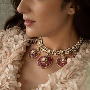 May include: A woman wearing a pink and white ruffled top with a gold and ruby necklace. The necklace has a large central pendant with smaller pendants on either side.