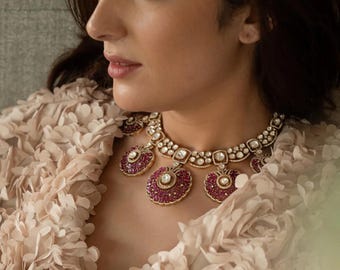 Puede incluir: Una mujer lleva una blusa de color rosa y blanco con volantes y un collar de oro y rubíes. El collar tiene un gran colgante central con colgantes más pequeños a cada lado.