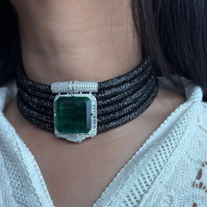 Puede incluir: Un collar gargantilla negro con múltiples hilos, con un gran colgante rectangular de esmeralda verde. El colgante está rodeado por un halo de piedras blancas. El collar es una pieza llamativa.