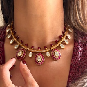 May include: A gold necklace featuring a red and white stone design. The necklace has a central pendant with a large white stone surrounded by smaller red stones. The pendant is flanked by two smaller pendants with similar designs.