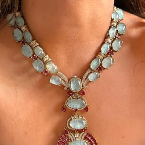 Puede incluir: Un collar de varias hileras con piedras cabujón ovaladas de color azul claro y pequeñas gemas rojas engastadas en un marco metálico. El diseño incluye un colgante central con una piedra azul grande rodeada de piedras rojas y detalles.