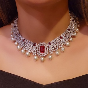 Puede incluir: Un collar de plata y diamantes blancos con una gran piedra roja en el centro. El collar tiene una fila de perlas que cuelgan de la parte inferior.