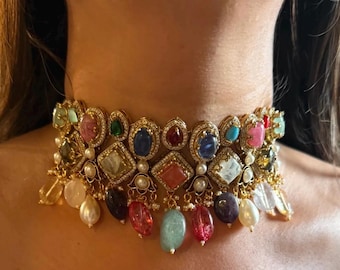May include: A gold-toned choker necklace featuring an array of colorful gemstones in various shapes. The necklace includes emerald green, sapphire blue, ruby red, and other vibrant stones, accented with small pearls and dangling teardrop-shaped gems.