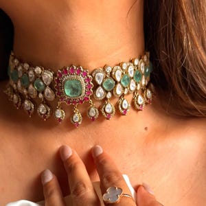 May include: A gold-toned choker necklace with emerald and ruby accents. The necklace features a central square emerald surrounded by red stones, with teardrop-shaped pendants. The design includes clear, round stones and a floral pattern.