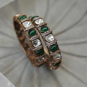 May include: Two gold-toned bangles with alternating emerald green and clear rectangular gemstones. The bangles are adorned with small, sparkling accents, creating a luxurious and elegant appearance. The jewelry is displayed on a soft, light-colored surface.