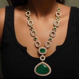 May include: A gold necklace with a large emerald green pendant. The pendant is surrounded by smaller diamonds. The necklace is made of gold and has a chain of oval links.