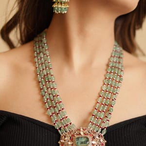 Puede incluir: Un elegante conjunto de collar y pendientes. El collar presenta múltiples hilos de cuentas verde claro intercaladas con cuentas rojas, que culminan en un gran colgante con una piedra central verde y detalles ornamentados. Los pendientes reflejan el diseño del colgante.