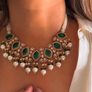 May include: A gold-toned necklace featuring large, oval emerald-green stones and smaller clear stones. The necklace has a pearl strand and dangling pearl accents. The necklace is worn around the neck.