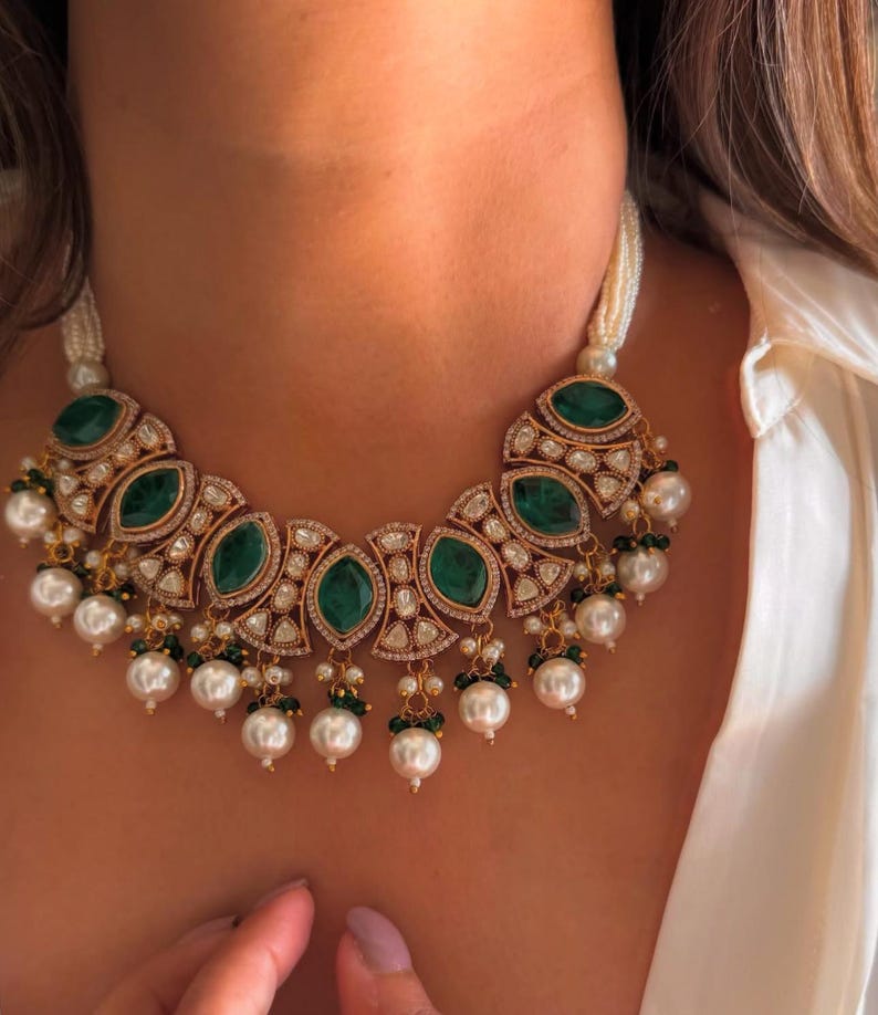 May include: A gold-toned necklace featuring large, oval emerald-green stones and smaller clear stones. The necklace has a pearl strand and dangling pearl accents. The necklace is worn around the neck.