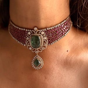 Może przedstawiać: Luksusowy naszyjnik typu choker z szerokim pasmem małych, czerwonych kamieni szlachetnych i centralną zawieszką. Zawieszka prezentuje duży, kwadratowy zielony kamień szlachetny i zielony kamień szlachetny w kształcie łzy, oba otoczone mniejszymi, przezroczystymi kamieniami.