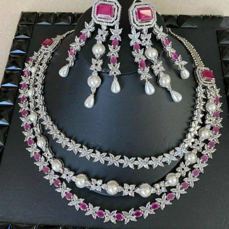 K&ouml;nnte beinhalten: Ein dreireihiges Halsketten- und Ohrring-Set mit silberner Oberfl&auml;che. Die Halskette hat ein Design aus abwechselnden klaren Zirkonia-Steinen und rosa ovalen Steinen. Die Ohrringe sind H&auml;ngeohrringe mit einem &auml;hnlichen Design.