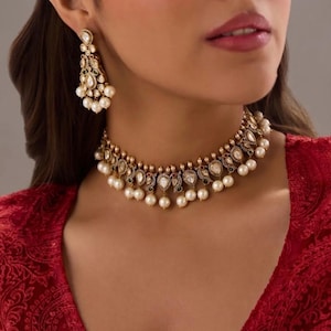 Könnte beinhalten: Ein goldfarbenes Halsketten- und Ohrring-Set. Die Halskette und die Ohrringe sind mit weißen Perlen, klaren Edelsteinen und farbigen Akzenten versehen. Der Schmuck wird zu einem roten Kleidungsstück getragen.