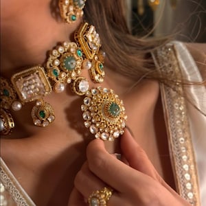 May include: A detailed gold-coloured jewellery set, including a necklace, earrings, and ring. The necklace and earrings feature emerald green and clear stone details, with pearl-like elements. The ring has a matching design.