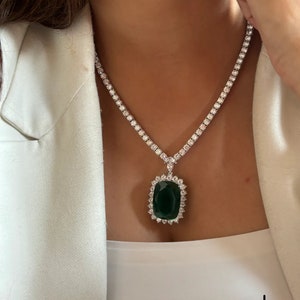 Puede incluir: Un collar de plata con un gran colgante de piedra preciosa verde esmeralda rodeado de pequeñas piedras preciosas transparentes. El collar es de estilo cadena de tenis.