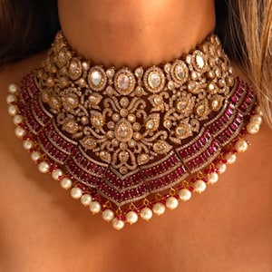 Peut inclure: Un collier doré orné de détails complexes. Le collier présente des pierres claires de forme ovale et de petits accents scintillants. Une section de pierres rouge foncé est située en dessous, avec une bordure de petites perles blanches.