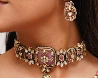 Pode incluir: Um colar choker dourado com uma pedra rosa no centro e detalhes florais. O colar tem um pingente pendurado com uma pedra grande e transparente. O colar é combinado com brincos combinando com pedras rosa e detalhes transparentes.