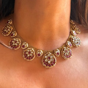 May include: Gold-toned necklace featuring a series of circular and teardrop-shaped pendants. The pendants are adorned with red and white gemstones, creating a detailed and ornate design. The necklace is worn around the neck.