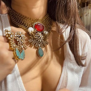 May include: A gold-toned necklace with a red oval gemstone and a teardrop-shaped turquoise gemstone pendant. The necklace features a chain design and is paired with matching earrings with turquoise and clear gemstones. The jewelry is ornate and detailed.