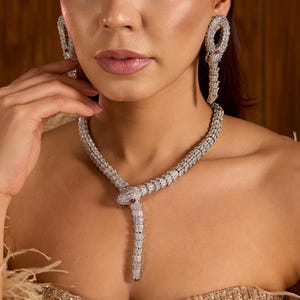May include: A silver-toned snake-shaped necklace and matching drop earrings, both encrusted with clear pavé stones. The snake head features small red eyes, and the body of the necklace wraps around the neck with a flexible, segmented design.