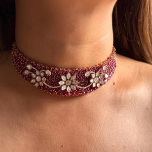 Puede incluir: Un collar gargantilla rojo rubí con un diseño floral. El collar presenta un patrón de pequeñas piedras rojas redondas y detalles florales blancos. El collar se lleva alrededor del cuello.