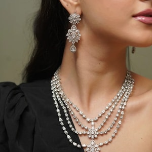 Può includere: Una collana d'argento con una catena multistrato e un grande ciondolo decorato. Il ciondolo è fatto di pietre trasparenti e ha una forma a goccia. La collana è indossata da una persona con i capelli scuri.