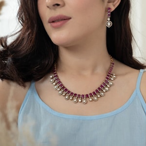 Op de afbeelding: Een gouden ketting met een rij roze edelstenen en hangende peervormige accenten. De ketting wordt gedragen door een persoon met lang donker haar.