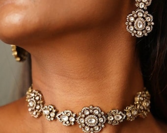 Könnte beinhalten: Eine goldene Choker-Kette mit einem zarten Design, das mit mehreren weißen Edelsteinen besetzt ist. Die Kette ist aus Metall und hat ein florales Muster.