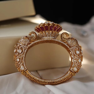 Puede incluir: Un brazalete de oro con detalles intrincados, con dos figuras de elefantes a cada lado y una corona de joyas roja y blanca en el centro.