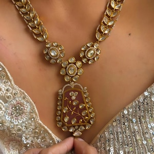 Puede incluir: Un collar de oro con un gran colgante de piedra preciosa rosa. El colgante está rodeado de piedras preciosas más pequeñas y transparentes y tiene un diseño floral. El collar está hecho de una cadena de hojas de oro con piedras preciosas transparentes.
