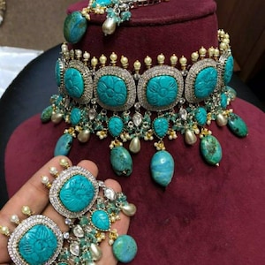 May include: A turquoise jewelry set featuring a necklace, bracelet, and earrings. The necklace has large, square turquoise stones with floral carvings, surrounded by silver and small pearls. The earrings mirror the necklace design, with additional turquoise drops.