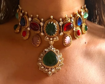 May include: A gold-toned statement necklace featuring a variety of colorful gemstones. The necklace has a central pendant with a large green stone, surrounded by smaller gems in red, blue, and pink. The design includes intricate detailing and small pearl accents.